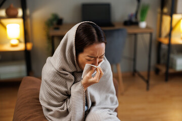 Sick Asian woman blowing nose with tissue wrapped in warm blanket