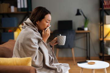 Side view of sick Asian woman blowing nose and holding mug on couch