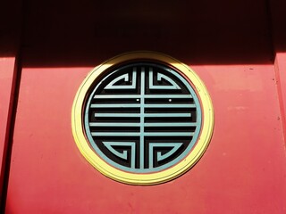 A centered close-up of a circular window with teal and yellow geometric latticework set into a vibrant red wall, featuring high-contrast shadows from direct sunlight.
