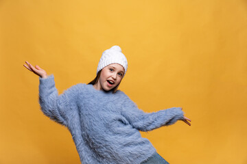 Happy child girl in blue sweatshirt and knitted hat with pompom dancing on yellow background. The concept of children emotions, joy and delight. Advertising clothing and sales. Ski resort, copy space