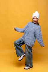 Happy child girl in blue sweatshirt and knitted hat with pompom dancing on yellow background. The concept of children emotions, joy and delight. Advertising clothing and sales. Ski resort, copy space