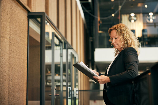 Mature businesswoman reading document while standing in law firm corridor