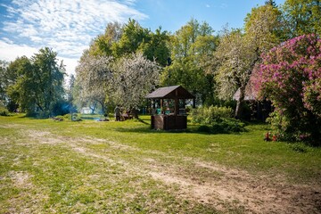 Obraz premium A wide grassy yard with a hand pumped wooden well and a steep roof barn. Two people tend a campfire by a picnic table under white blossoms in bright morning light.