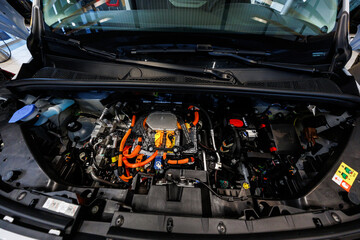Obraz premium Open hood view shows orange high voltage cables, sealed inverter unit, coolant reservoirs, and a 12V battery indoors under showroom lighting during daytime.