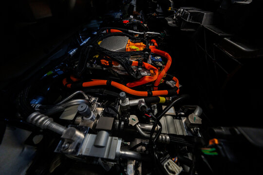 Close up of an electric vehicle powertrain shows orange high voltage cables to inverter and power electronics, with metal coolant lines in low light workshop setting.