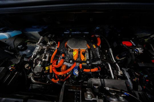 Close up under a car hood shows an electric powertrain with orange cables, sealed inverter, braided harness, coolant lines, and warning labels in workshop daylight.