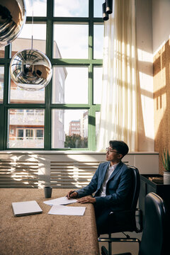 Thoughtful male business professional sitting at table with agreement papers in office