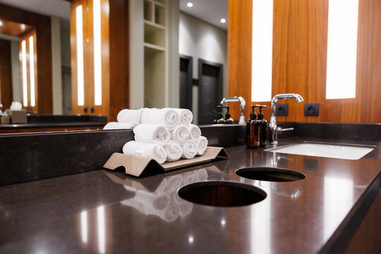 A dark stone vanity holds rolled white towels, chrome faucets, amber soap dispensers, and built in waste chutes, with warm wood walls and tall light panels in a mirror.