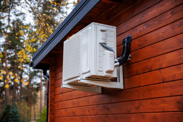 A white split system outdoor unit sits on metal brackets against reddish timber siding, with insulated lines in a black conduit on a cool day with soft diffused light.