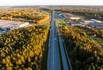 Aerial view of divided highway with warehouses and loading bays. Late day light casts long shadows on lanes and signage as trucks and cars move past conifer stands and frontage road.