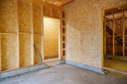 Workers build interior rooms with OSB on stud walls, exposed joists, and a raw concrete slab. Open doorways lead to a corridor and stairwell during mid construction.