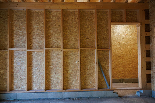 An unfinished interior shows vertical 2x4 studs, OSB sheathing, and a rough doorway on a concrete block stem wall. Warm tungsten light spills in, highlighting a leaning conduit pipe.