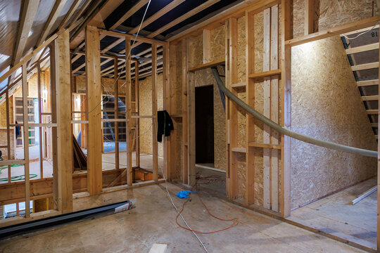 Interior attic renovation shows stud framing, OSB sheathing, rafters, wires, and a large flexible duct  tools and cords indicate active work under even work lighting.