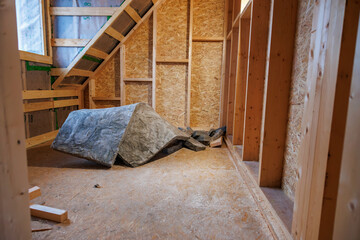 An unfinished attic shows pine studs, OSB, a taped window opening, and a torn roll of mineral wool...