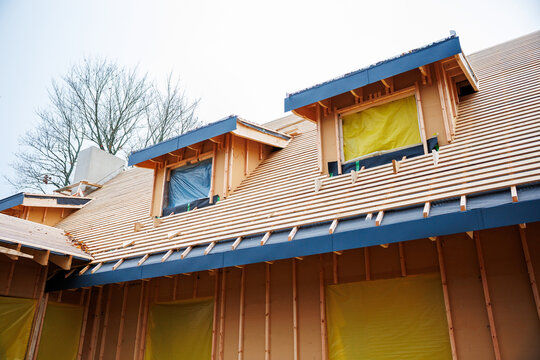 Contractors build a multi dormer steep pitched roof with battens, sealed plastic windows, extended eaves, and blue flashing in overcast light during a cool season.