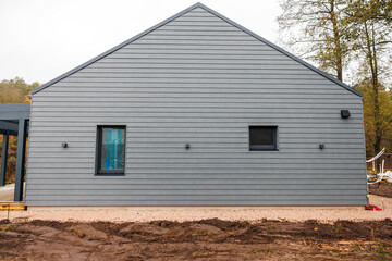 Obraz premium Modern single story house with sharp gable and gray cladding, front on and symmetrical, late autumn, two windows, four wall lights, gravel edge, bare soil, tall trees