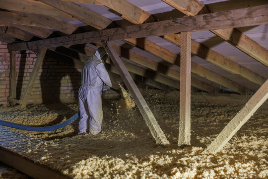 An installer in a white suit blows loose fill insulation through a hose in a low timber framed attic with a rough brick gable wall and exposed rafters during renovation.