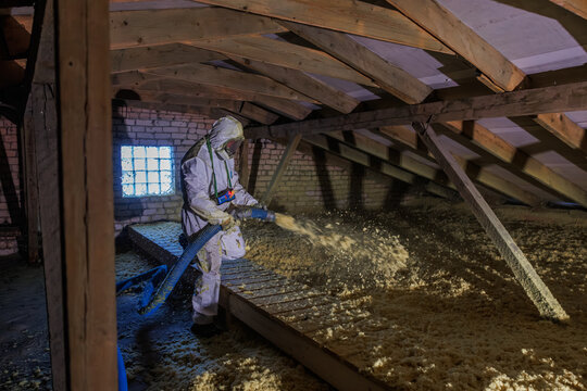 Worker in protective suit uses blue hose to spread loose fill insulation in a timber framed attic with exposed rafters and a glass block window under harsh side light.