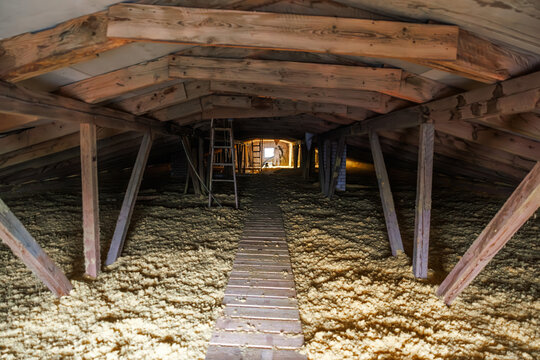 A tunnel like attic shows rough sawn rafters, diagonal braces, and a narrow wooden catwalk over loose fill insulation. A lit hatch, wiring, and a small gable vent appear.