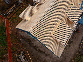 Aerial view shows a steep gable roof under construction with battens, dark underlayment, framed dormer openings, fallen leaves, ladders, stacked lumber, and a safety fence.
