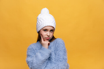 Close up studio portrait of pensive little girl holding index finger near her mouth, wearing in blue sweater, white knitted hat on yellow background. Copy space. High quality photo