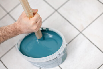 Male hand stirring blue paint in a bucket with a wooden stick, showcasing the preparation process for home improvement and DIY projects with vibrant color