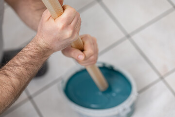Male painter stirring teal paint in a bucket with a wooden stick, demonstrating preparation for home improvement project, showcasing craftsmanship and dedication to quality work