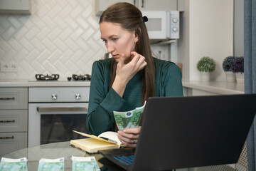 Worried freelance woman counting euro banknotes in her home kitchen using laptop and notebook,...