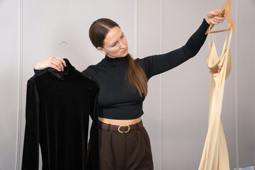 Professional fashion designer holding two dresses on hangers, comparing a black velvet dress and a gold silk one while deciding which one is best for her new collection