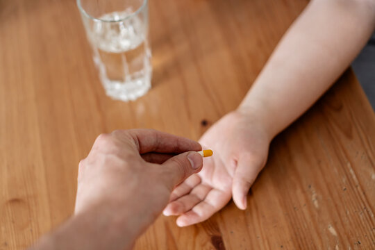 Parent giving prescribed pill to his sick shild at home. Personal perspective photo of father's hand with capsule and little boy hand with glass of water on the table to wash down the medicine.