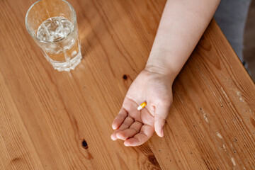 Kid holding capsule in the palm of his hand over tabletop with glass of water on the table with copy space. Concept of home treatment.