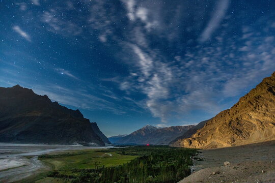 View of the sprawling green valley and winding river nestled between towering mountains under a dramatic sky streaked with clouds and stars, Shigar, Gilgit Baltistan, Pakistan.