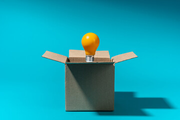 Minimalist photo of opened cardboard box with bulb inside over turquoise blue background, harsh shadows. Thinking outside the box, great idea and creative problem solving concept with copy space.