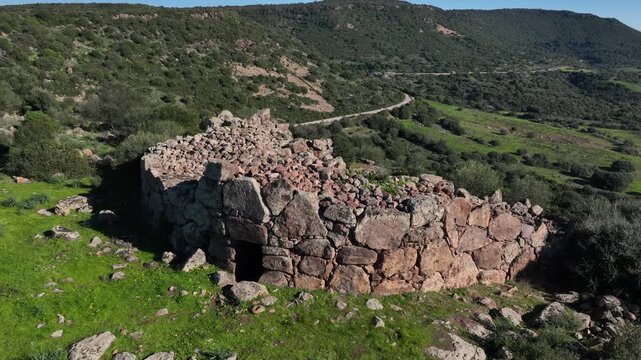 Aerial view of a Nuragic stone building ruins set against the backdrop of lush green hills creating a stark contrast with the rugged, ancient structure, Olmedo, Sardinia, Italy.