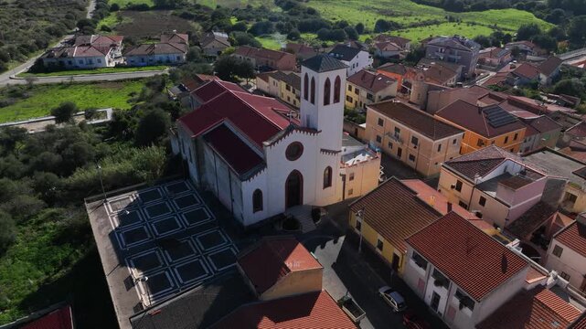 Aerial view of a white church with a red roof standing tall among the buildings of the town with its unique architecture, Olmedo, Sardinia, Italy.