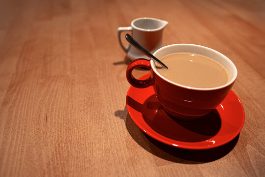 A red ceramic cup filled with coffee and milk sits on a wooden table next to a small white creamer pitcher.