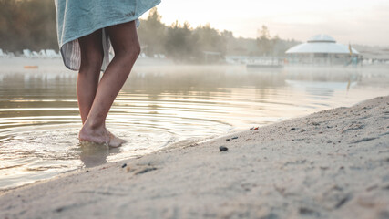 Person's bare feet wading into calm lake water at sunrise with morning mist