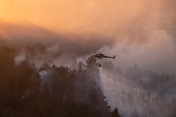 Wildfire suppression helicopter with yellow helibucket releasing water onto a forest fire in Ourense