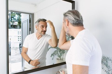 Obraz premium A mature man checks his hairstyle in the mirror at home, touching his gray hair with confidence. Wearing a white shirt, he shows self care, grooming routine, and a calm lifestyle moment.