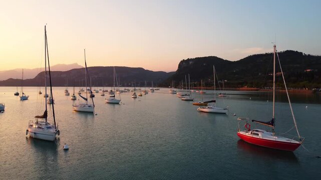 Drone fly-through of white sailing yachts anchored in perfect geometric rows on Lake Garda, Italy, under a vibrant sunset, with surrounding mountains reflecting on calm water, showcasing symmetry 