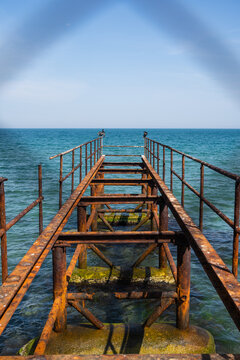 View of a weathered pier, its rusty metal framework contrasting with the turquoise sea and clear blue sky, extending into the horizon, Sozopol, Burgas, Bulgaria.