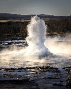 View of a powerful geyser erupting with a burst of water and steam against a backdrop of brown fields and distant mountains, Iceland.
