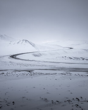 View of a sinuous black road cutting through the expansive white canvas of snow-covered fields and distant mountains under a pale sky, Hverir, Iceland.