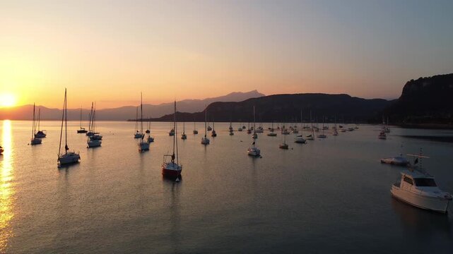 drone view of white sailing yachts anchored in perfect geometric rows on Lake Garda, Italy, under a vivid sunset, reflecting golden light on calm water, evoking elegance and serenity.