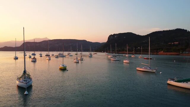 Drone fly-through of white sailing yachts anchored in perfect geometric rows on Lake Garda, Italy, under a vibrant sunset, with surrounding mountains reflecting on calm water, showcasing symmetry 