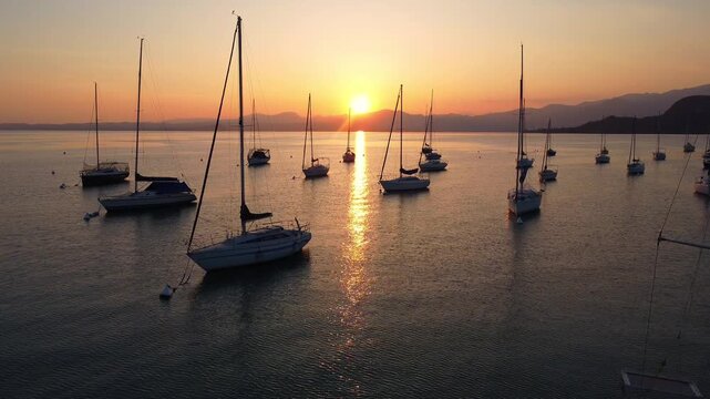 Drone fly-through of white sailing yachts anchored in perfect geometric rows on Lake Garda, Italy, under a vibrant sunset, with surrounding mountains reflecting on calm water, showcasing symmetry 