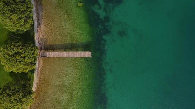 Top-down drone video of a geometric wooden pier extending into Lake Garda with clear green water, surrounded by lush trees, capturing symmetry, nature, and serene lakeside scenery