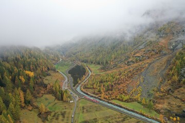 Aerial view of a train snaking through a valley, flanked by autumn-gold forests and a tranquil lake, all beneath a shroud of ethereal mist, Zermatt, Valais, Switzerland.