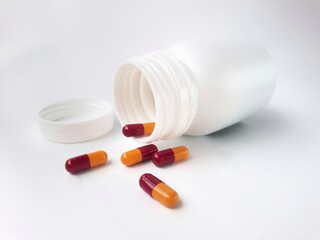 White plastic bottle with a screw-off cap on a plain background. Nearby, five capsules are scattered, each half orange and half red, suggesting medication or supplements