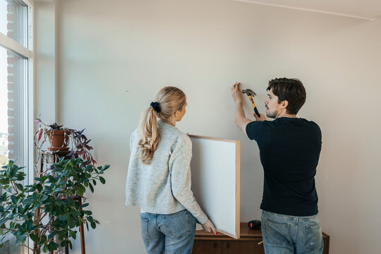 Young man hammering nail on wall while standing beside girlfriend holding picture frame